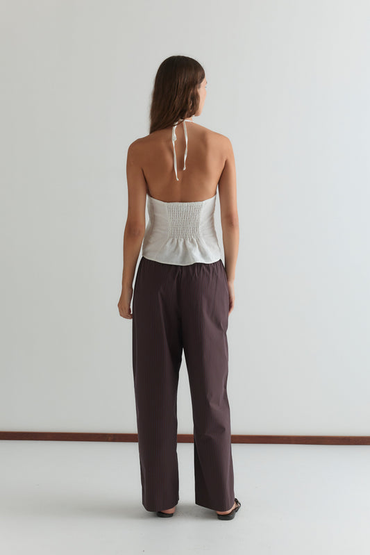 A woman stands facing away from the camera, wearing a white strapless Deiji Studios top and the Double Button Pant - Brown Stripe in a minimal, light-colored room.