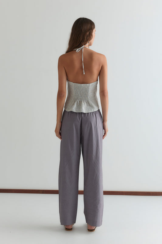 A woman stands facing away from the camera in a light gray halter top and Deiji Studios' Ease Trouser in Currant, shown in a minimal, light-filled room with a white wall and brown floor trim.