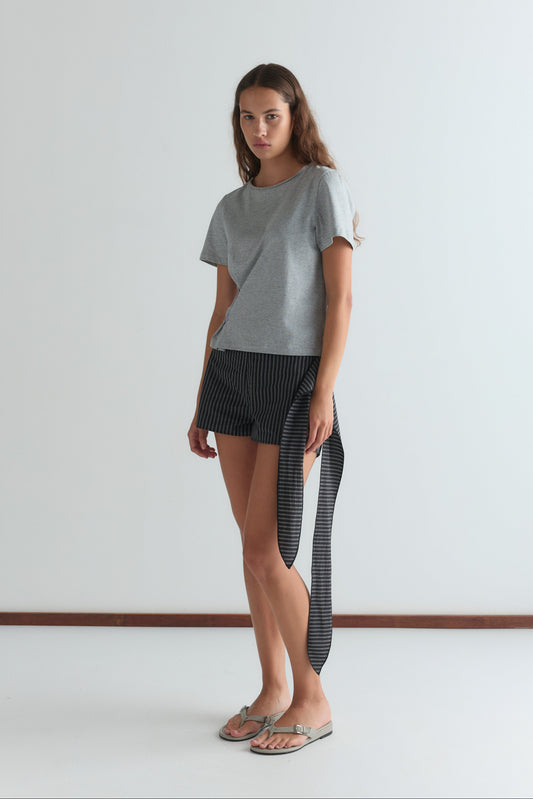 A woman with long brown hair stands indoors against a plain white wall, wearing a gray t-shirt, Deiji Studios Belted Short - Mono Stripe with a long side tie, and beige flip-flops.