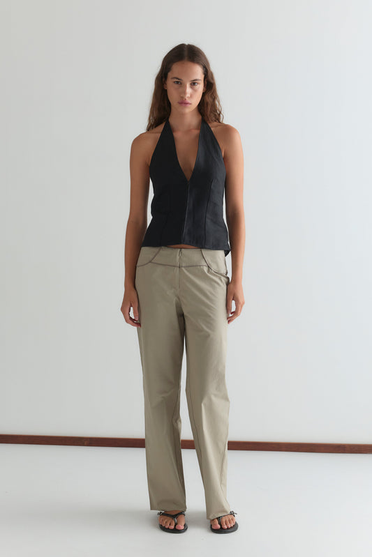 A woman with long brown hair and a neutral expression stands against a white wall, wearing the Close Halter Top - Black from Deiji Studios, beige zip-detail trousers, and black sandals.