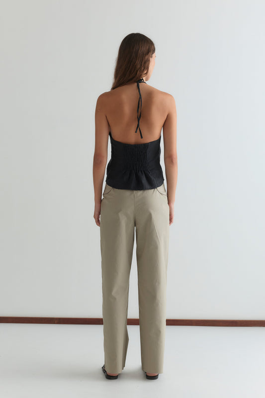 A woman faces away from the camera, wearing the Close Halter Top - Black by Deiji Studios with an open back and beige pants against a plain white background.
