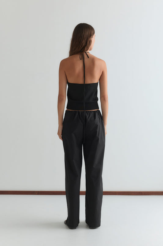 A person with long hair, facing away, wears the Seam Pant - Black and a black halter-top, standing against a plain white wall and floor.