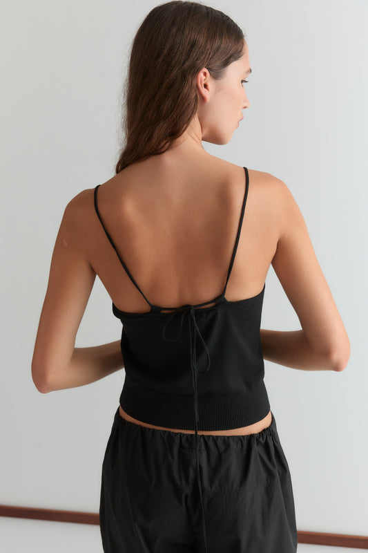 A woman with long brown hair faces away, wearing the Deiji Studios Drawcord Knit Top - Black with an adjustable ruched neckline and open back, paired with black pants against a plain light background.