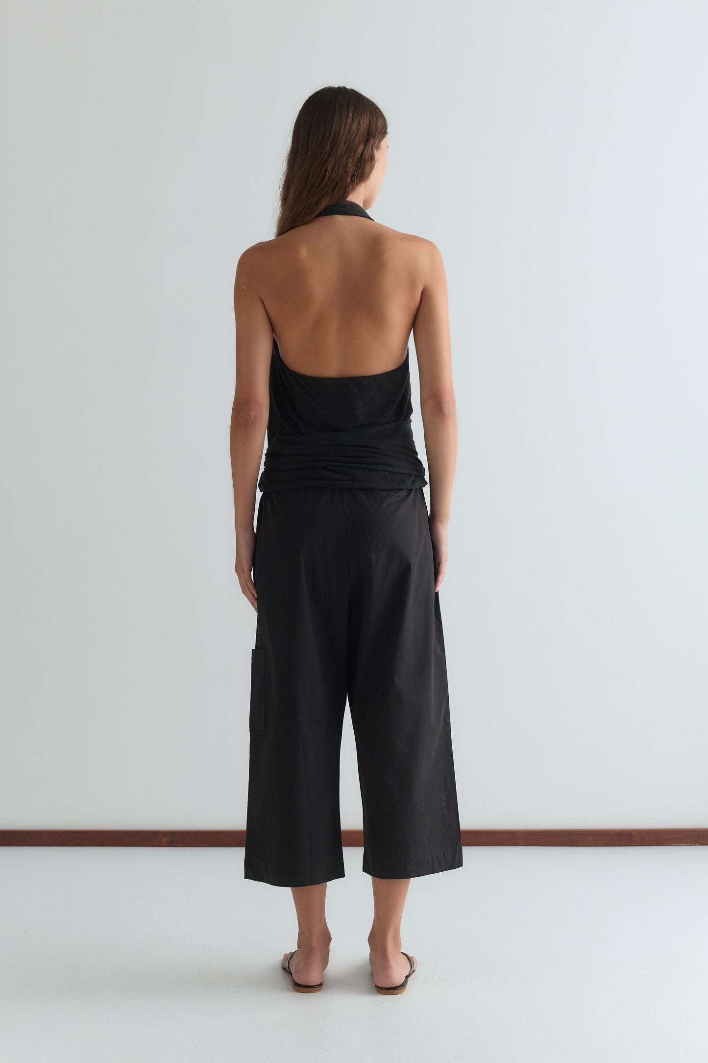 A person with long hair stands facing away, barefoot in Deiji Studios’ Mid Pocket Short - Black and a black halter top with an open back, against a plain white background.