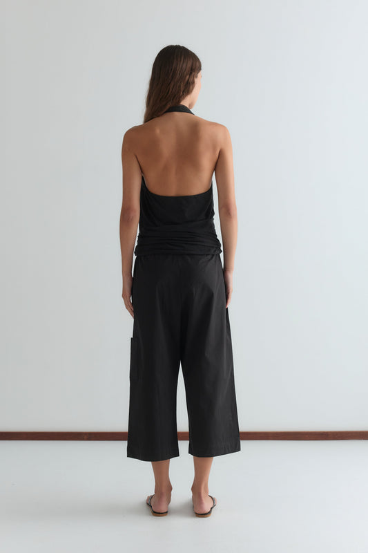 A person with long hair stands facing away, barefoot in Deiji Studios’ Mid Pocket Short - Black and a black halter top with an open back, against a plain white background.