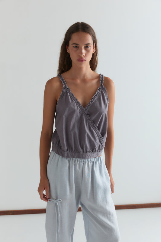A woman with long brown hair stands against a plain white background, wearing the Overlie Top - Currant by Deiji Studios and loose light blue pants, looking directly at the camera with a neutral expression.