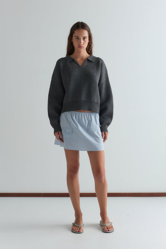 A woman with long brown hair stands against a plain white wall wearing a loose dark gray collared sweater, silver sandals, and the Pocket Skirt - Lake Blue Stripe by Deiji Studios. Her expression is neutral.