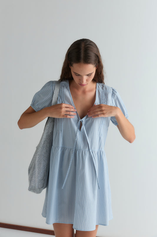 A woman with long hair stands against a plain background, looking down as she adjusts the front tie of her light blue, short-sleeved V Pleat Dress. She carries a textured gray tote bag on her shoulder.