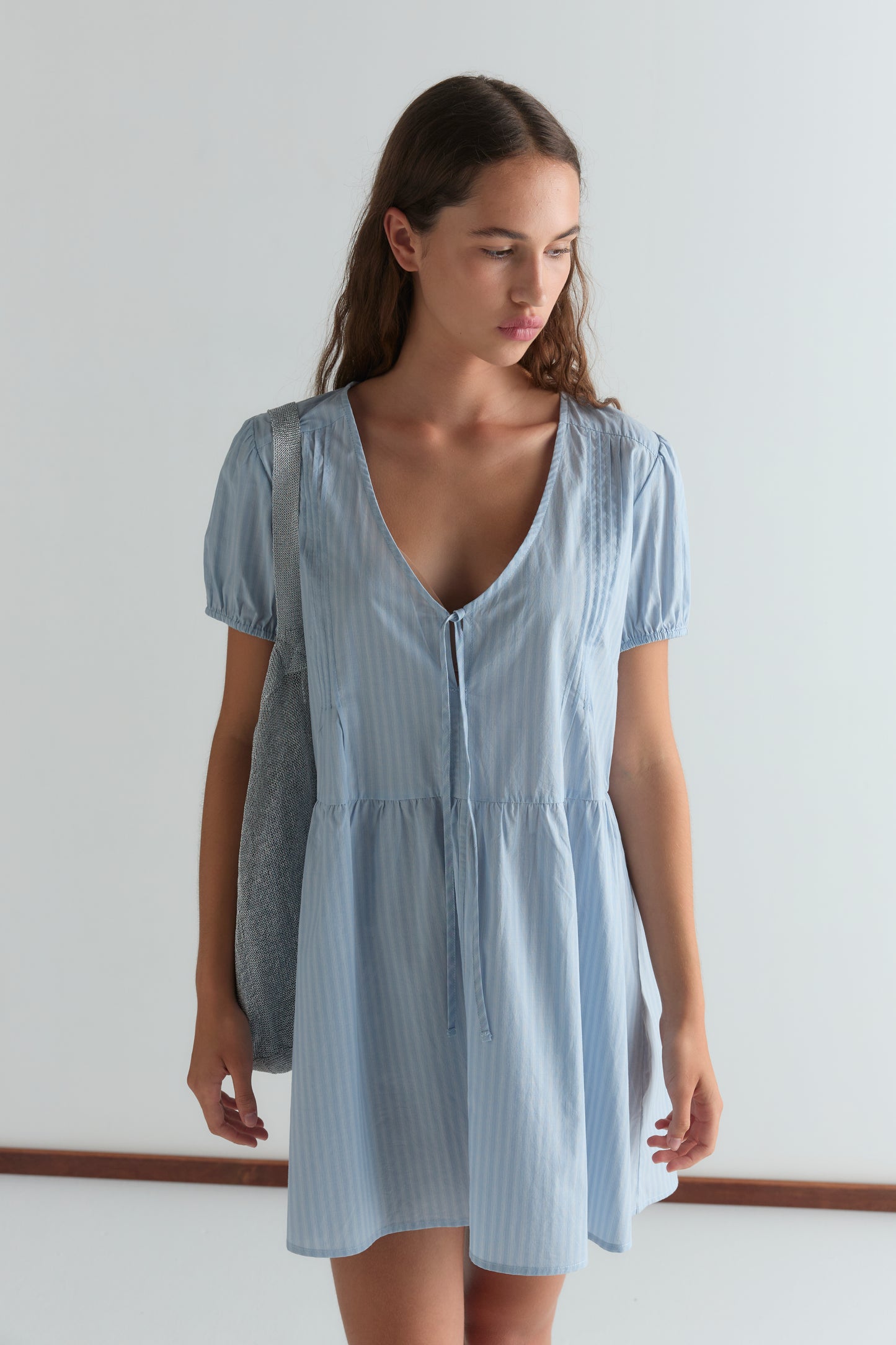 A young woman with long brown hair wears the V Pleat Dress - Lake Blue Stripe by Deiji Studios, standing indoors with a gray bag over her shoulder and looking down to her left against a plain white background.