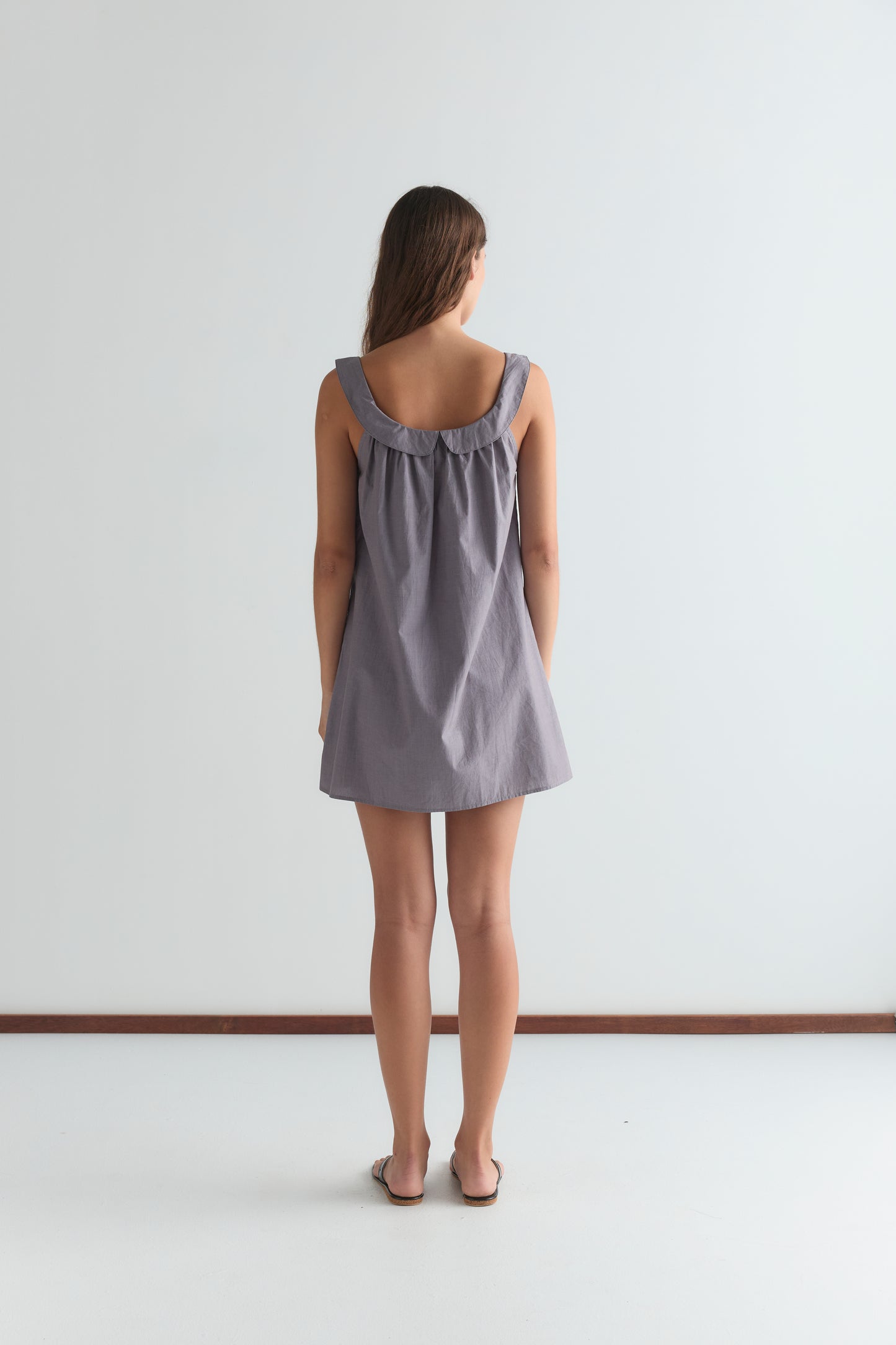 A woman faces away from the camera, wearing the Rounded Neck Dress - Currant, a loose sleeveless style with a gathered neckline, paired with brown sandals, against a plain white background.