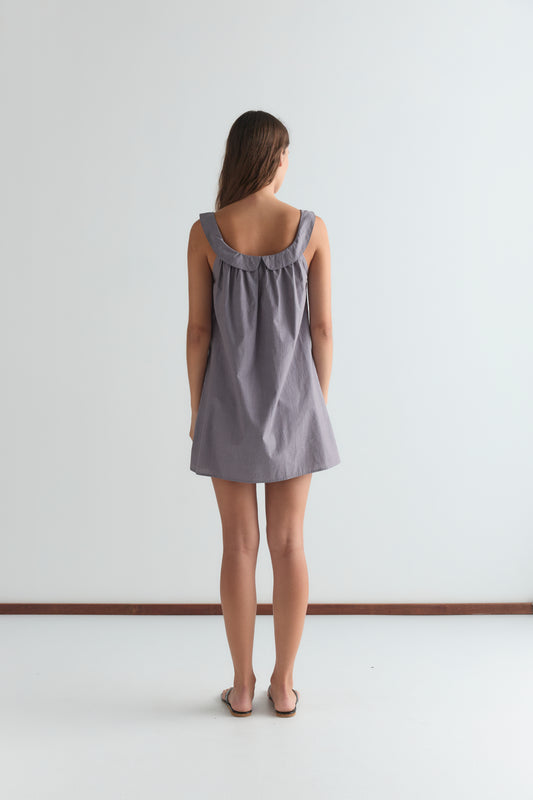 A woman faces away from the camera, wearing the Rounded Neck Dress - Currant, a loose sleeveless style with a gathered neckline, paired with brown sandals, against a plain white background.