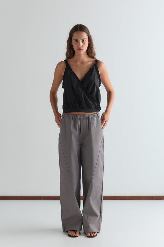 A woman stands against a plain white wall, wearing a sleeveless black wrap top and the Curve Pant in Lavender Smoke. The relaxed fit, mid-rise pants complement her look as she stands with hands in pockets, gazing directly at the camera.
