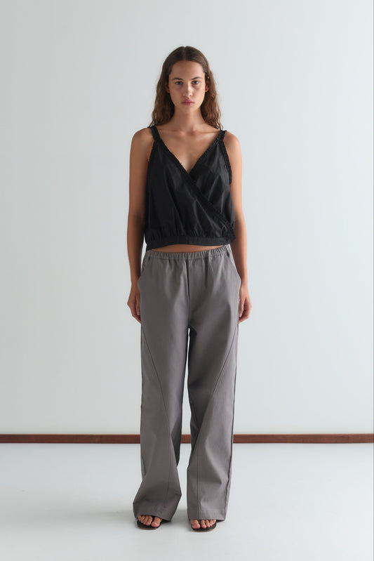 A woman faces forward in a sleeveless black wrap top and the Curve Pant - Lavender Smoke, featuring a relaxed fit and mid-rise waist, paired with black sandals against a plain white background.