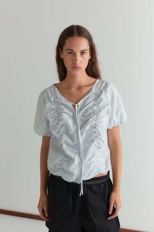 A woman with long brown hair wears the Crinkle Top - Powder Blue Stripe and black pants, standing against a plain white wall, looking directly at the camera with a neutral expression.