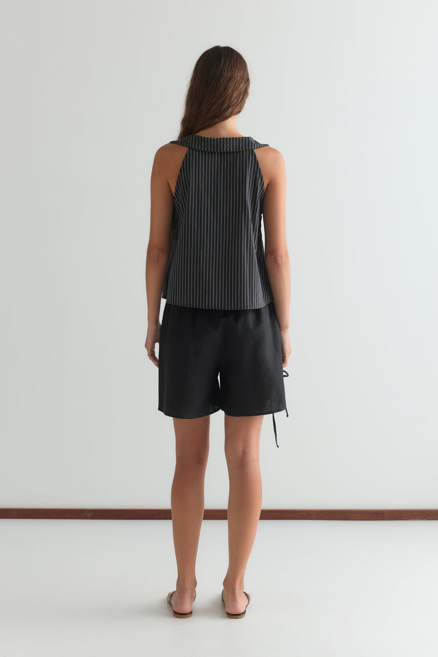 A woman stands facing away from the camera, wearing a sleeveless striped black top, the Eyelet Short - Black by Deiji Studios, and sandals against a plain white background.