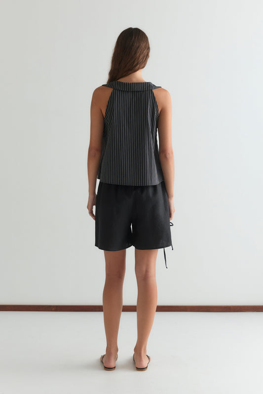 A woman stands facing away from the camera, wearing a sleeveless striped black top, the Eyelet Short - Black by Deiji Studios, and sandals against a plain white background.