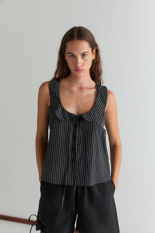A woman with long brown hair wears the Deiji Studios Round Collar Top - Mono Stripe, a sleeveless piece with black and gray stripes, rounded collar, and bow tie detail, paired with black shorts against a plain light background.