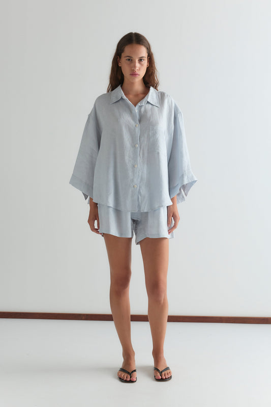 A person stands indoors against a plain white wall, wearing the 03 Set - Soft Blue from Deiji Studios, paired with black flip-flops.