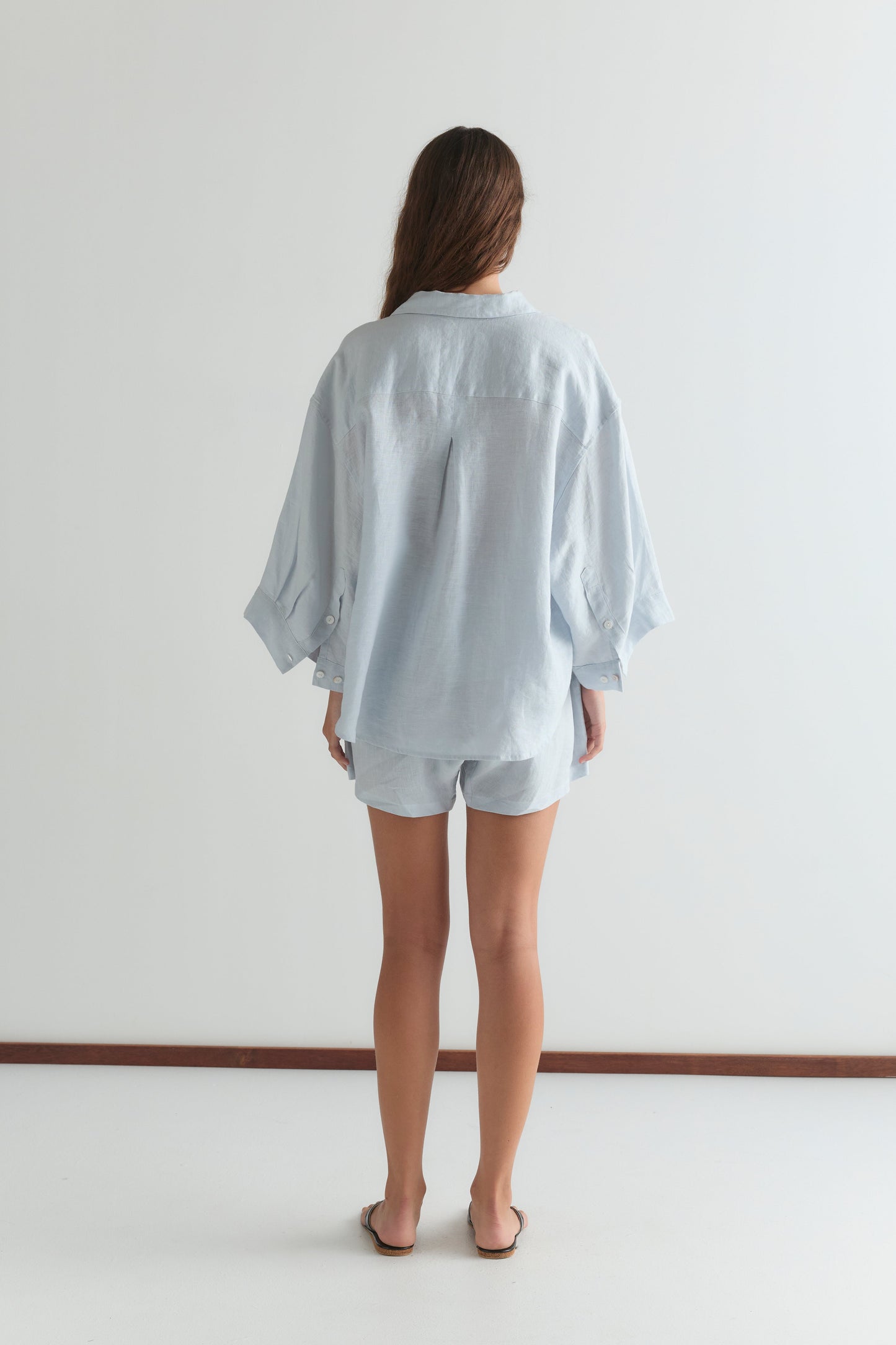 A woman, barefoot on a white floor against a plain white wall, wears the 03 Set - Soft Blue by Deiji Studios: an oversized linen shirt and matching boxer shorts.