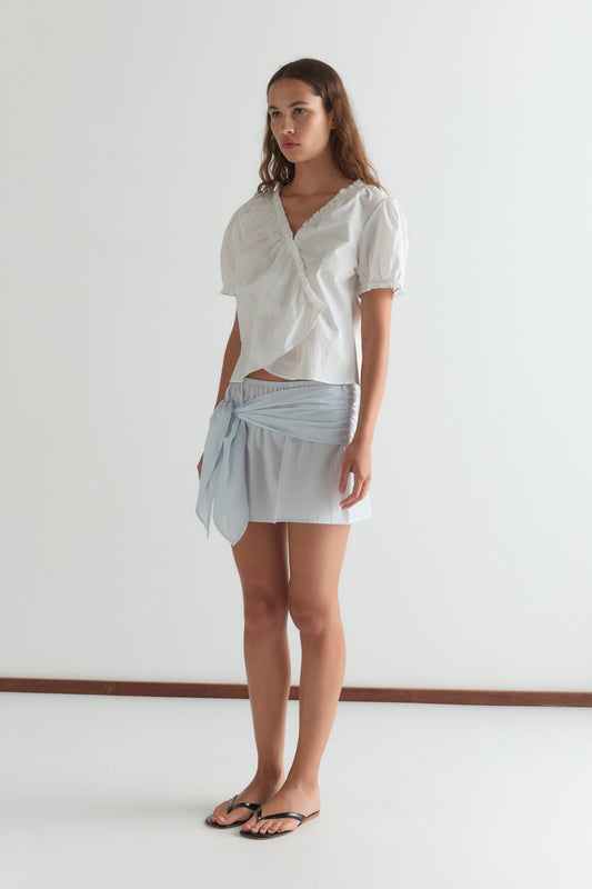 A woman stands indoors in a Cross Frill Top - White, paired with a light blue tie mini skirt and black flip-flops. She has long brown hair and a neutral expression, set against a plain light background.