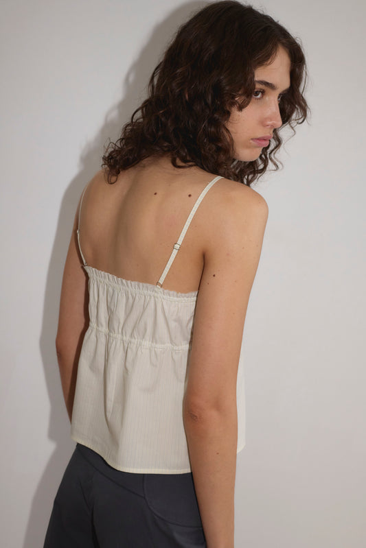 A person with curly brown hair wears the Tri Cami - Citrus Stripe by Deiji Studios with dark pants, standing against a plain white background and looking over their shoulder toward the camera.