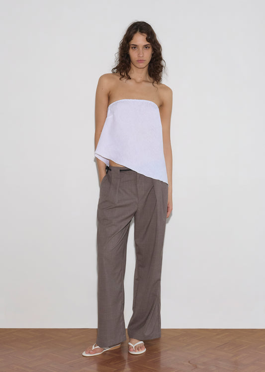 A person with wavy, shoulder-length brown hair wears a Deiji Studios Line Top - White with an asymmetric hem, loose brown trousers, and white flip-flops, standing against a plain white wall on a wooden floor.