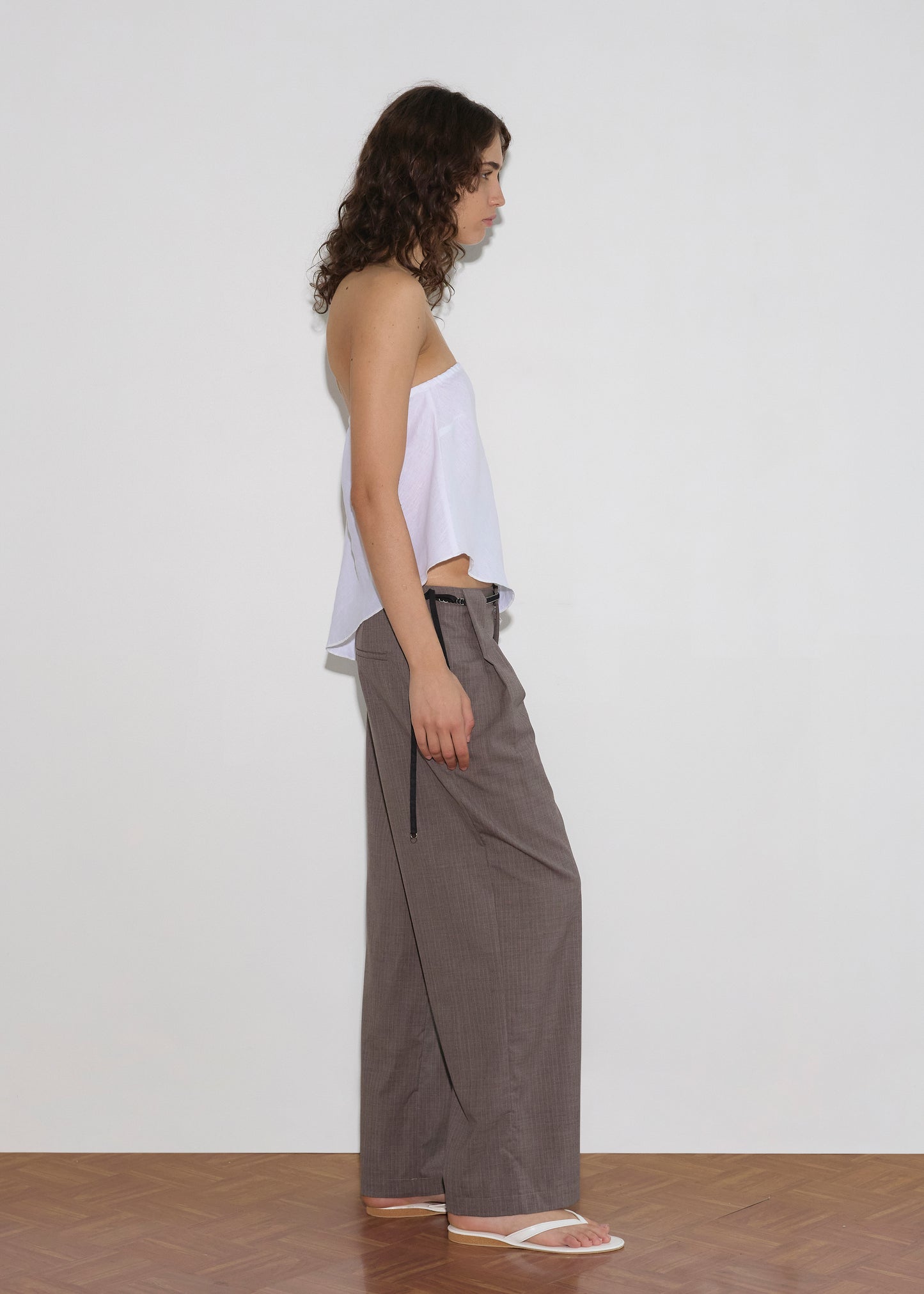 A person with curly brown hair stands in profile against a white wall, wearing a white strapless top, Belt Loop Pant - Grey Pink Stripe, and white sandals on a wooden floor.