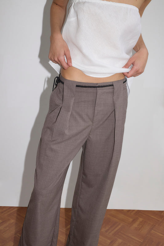 A person stands against a white wall, wearing a white strapless top and the Belt Loop Pant - Grey Pink Stripe, featuring relaxed wide legs, pleats, high waist, and black detailing at the waistband. Their face is not visible.