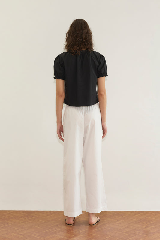 A person with wavy brown hair stands facing away, wearing the Cross Frill Top - Black with a cropped relaxed silhouette, paired with loose white pants and brown sandals on a wooden floor against a plain white wall.