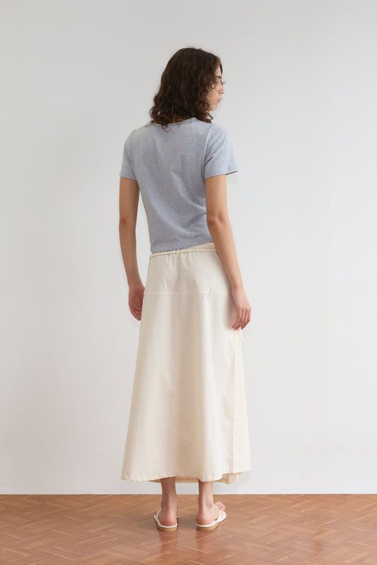 A woman stands indoors with her back to the camera, wearing a light gray short-sleeve t-shirt, the Loop Pocket Skirt - Off White by Deiji Studios, and white sandals on a wooden floor against a plain white wall.
