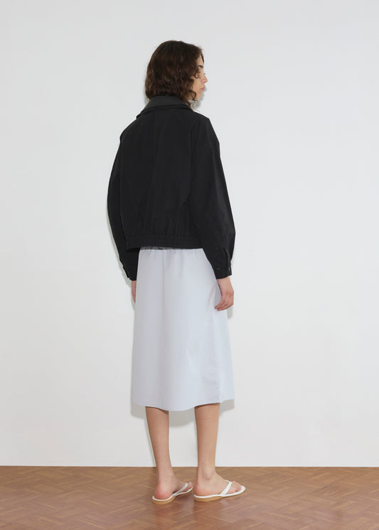 A person with curly hair faces a white wall, wearing the Collared Zip Jacket - Black Contrast, a light blue knee-length skirt, and white sandals on a parquet floor.