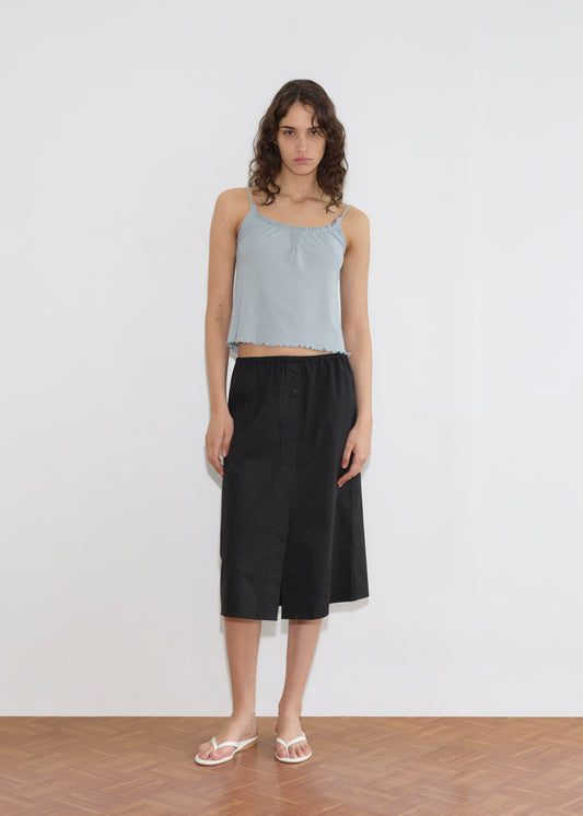 A woman with wavy brown hair wears the Cotton Flounce Tank - Baby Blue, paired with a black knee-length skirt and white flip-flops. She stands on a wooden floor against a plain white wall.