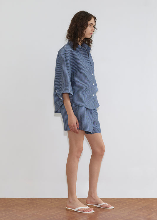 A person with wavy brown hair stands sideways in the Deiji Studios 03 Set - Azure Stripe, a loose linen shirt and matching boxer shorts, paired with white sandals, against a plain white wall on a wooden floor.