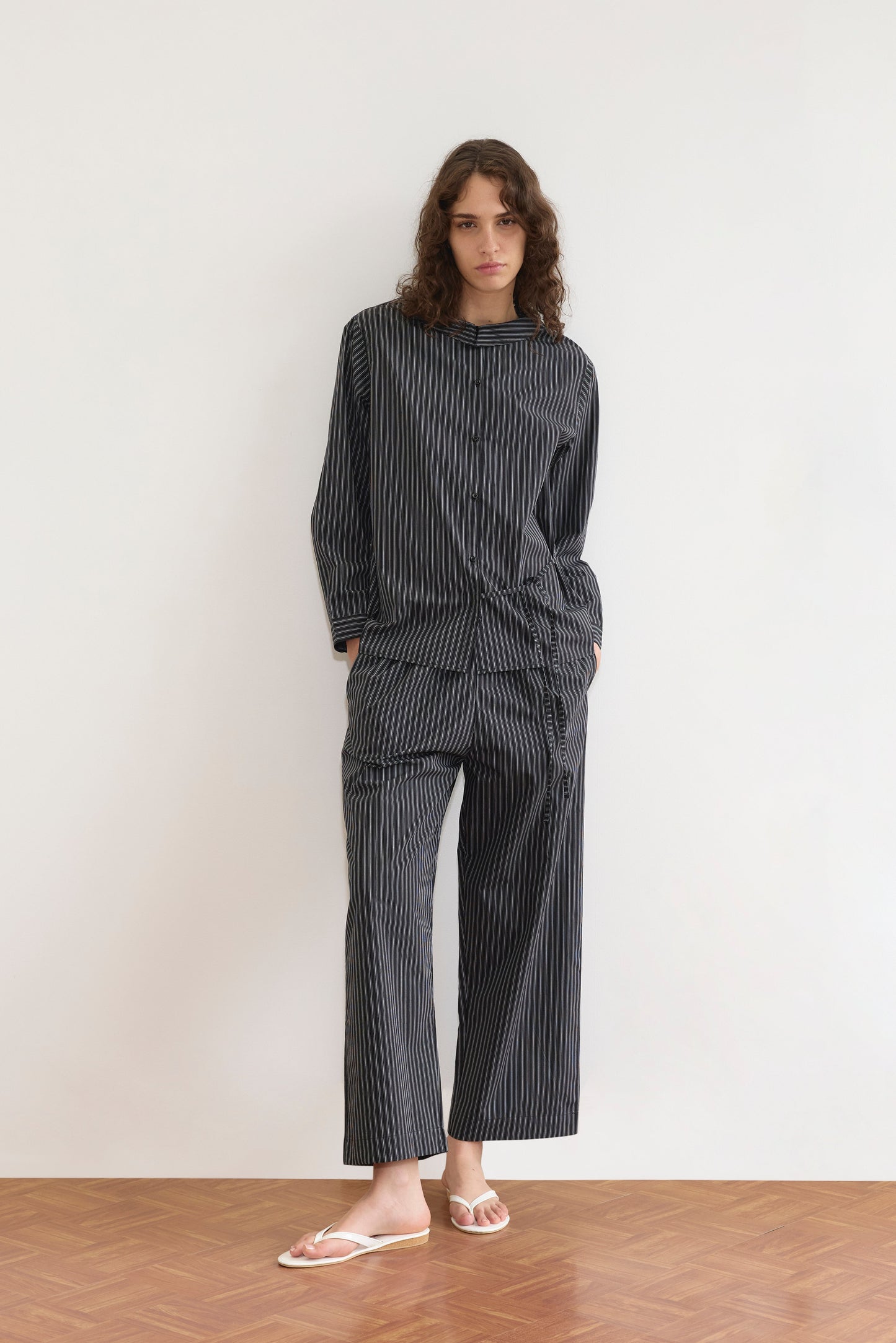 A person with long curly hair stands against a white wall, wearing the Deiji Studios Left Set - Mono Stripe: a lightweight, relaxed shirt and matching pants with hands in pockets, paired with white flip-flops on a wooden floor.