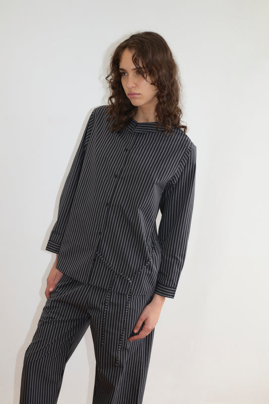 A person with wavy brown hair wears the Deiji Studios Left Set - Mono Stripe: a matching black and gray vertical striped lightweight shirt and pants, standing against a plain white background, looking slightly to the side with a neutral expression.