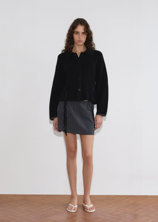 A person with long, curly hair wears the Tie Side Cardigan - Black, a short pinstriped skirt, and white sandals while standing against a plain white wall on a wooden floor.