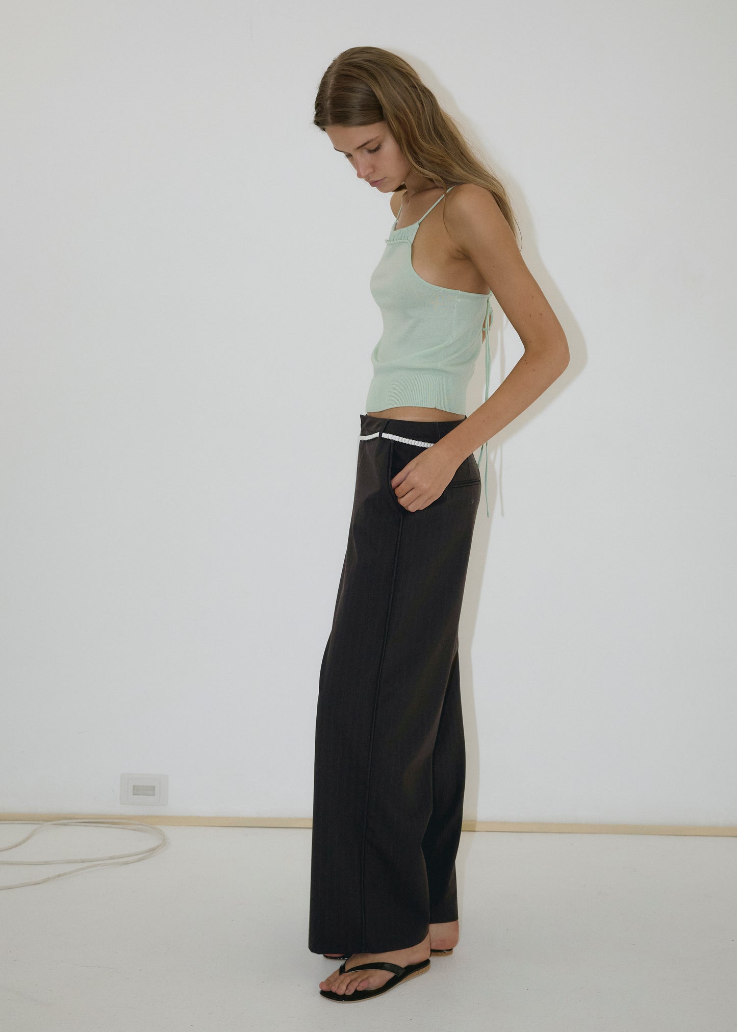 A woman stands sideways against a white wall, wearing a light green sleeveless top, the Bind Pant - Charred Black Stripe from Deiji Studios with a white belt, and black sandals. She looks down, arms relaxed by her sides.