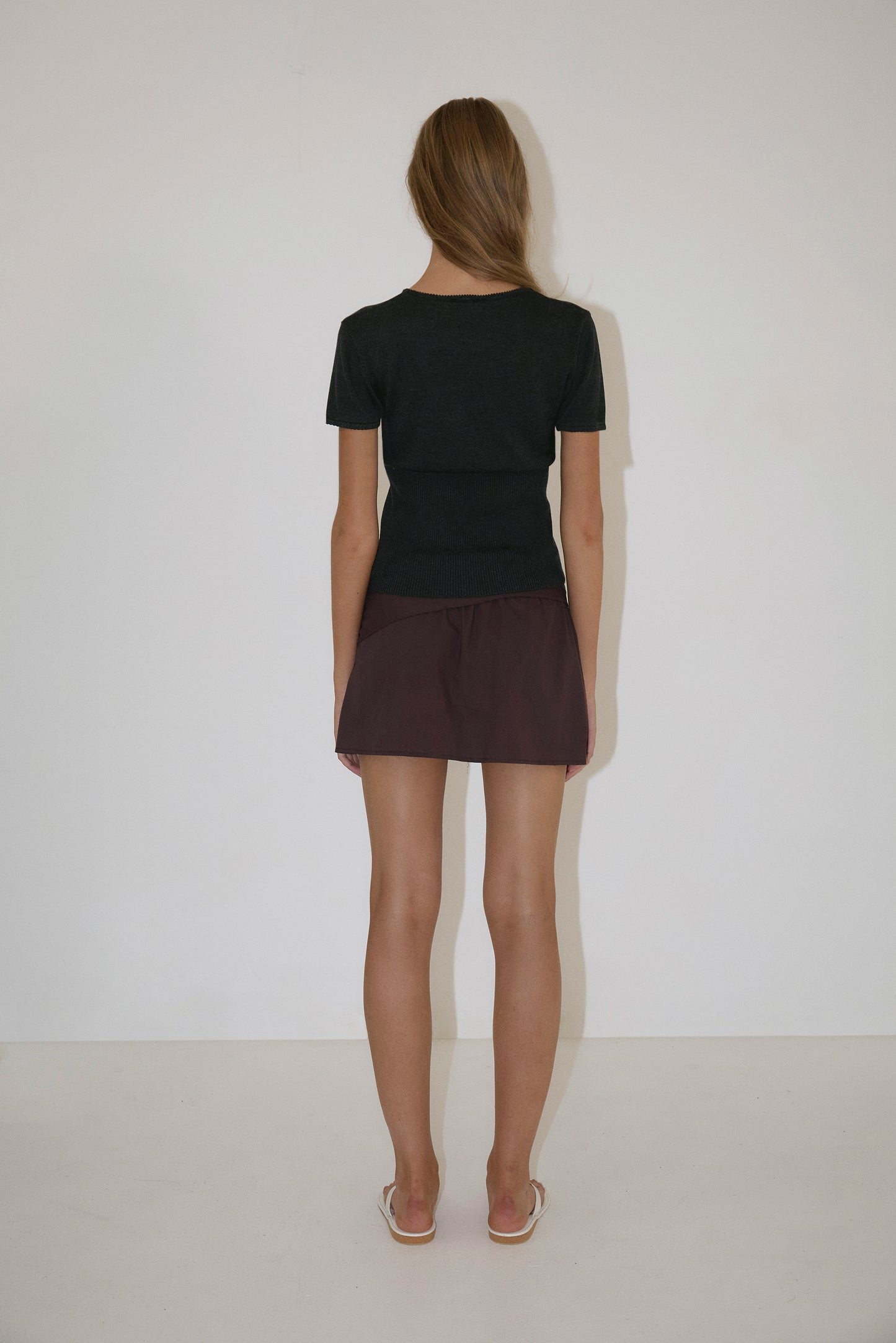 A woman with long hair faces away from the camera, wearing a Trim Knit Tee - Dark Grey by Deiji Studios, a dark brown mini skirt, and white flip-flops against a plain white wall and floor.