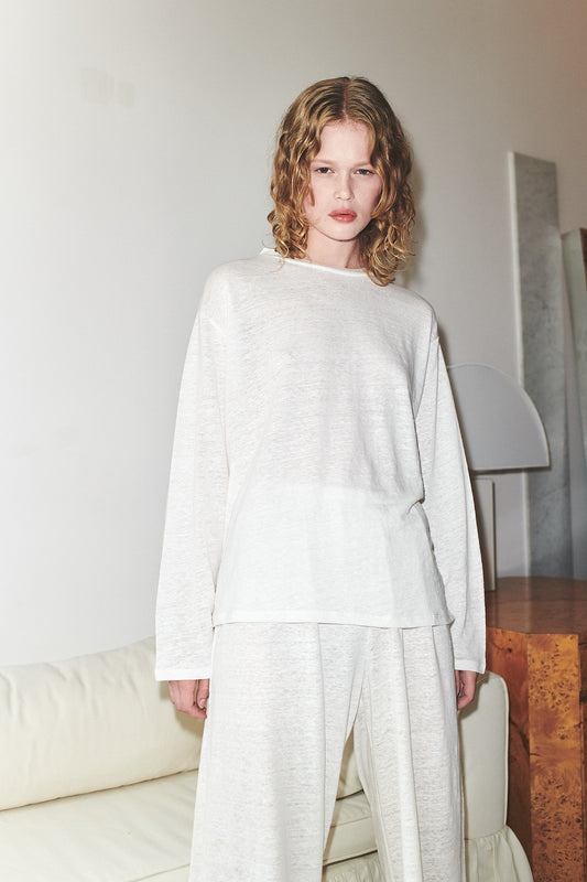 A person with wavy, shoulder-length blond hair wears the Deiji Studios soft long sleeve top in ecru, paired with matching wide-leg pants, standing indoors by a cream sofa and wooden side table for a relaxed loungewear look.