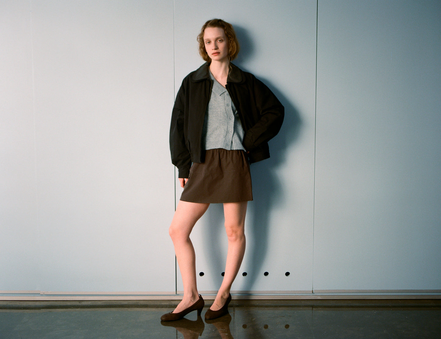 A person stands indoors by a plain light blue wall, wearing a black jacket, gray shirt, Deiji Studios Pocket Skirt - Sepia, and black flats. They have short wavy hair and stand relaxed with one hand on their hip.
