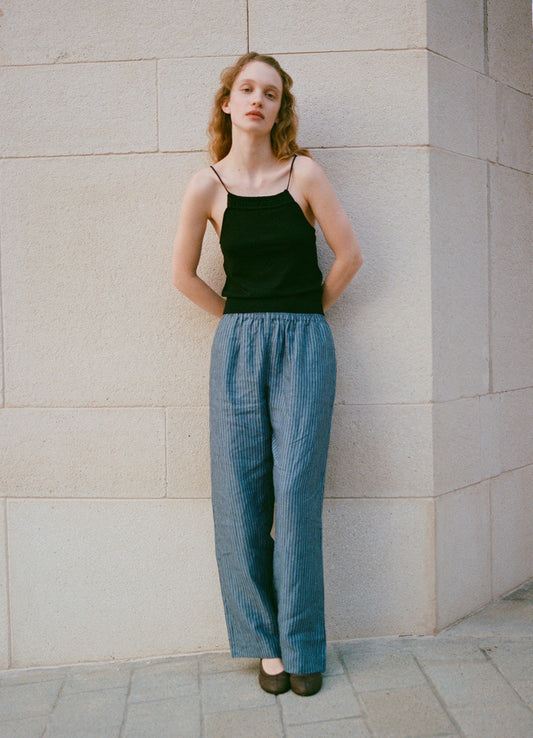 A woman with wavy hair stands against a light stone wall, wearing a black sleeveless top and the Linen Ease Trouser - Azure Stripe. Her arms rest behind her back as she gazes calmly at the camera, embodying relaxed, effortless style.