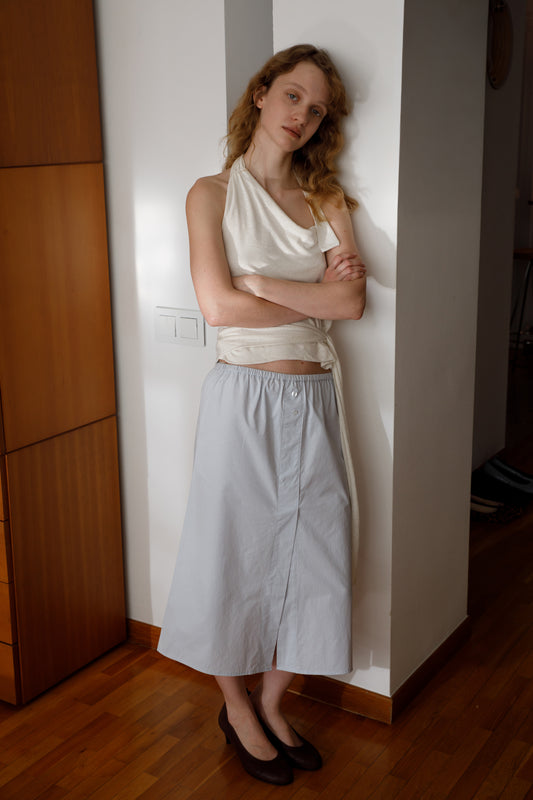 A woman with wavy blonde hair, wearing a sleeveless white top and the Overlap Skirt - Blue by Deiji Studios, stands indoors against a white wall with arms crossed, gazing calmly at the camera.