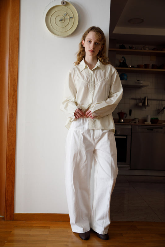 A young woman with long hair stands indoors near a kitchen, wearing a loose-fitting cream blouse and the Pleat Pant - White. She is by a white wall with a round clock above her, standing on wooden flooring.
