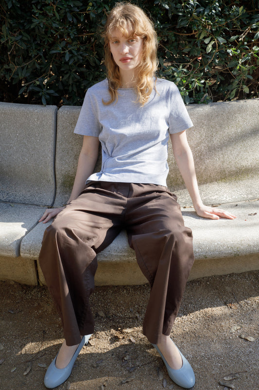 A person with wavy blond hair sits on a stone bench outdoors, wearing a light gray T-shirt and the Deiji Studios Contrast Waist Pant - Brown in a relaxed straight-leg fit. Light blue shoes and sunlit green bushes complete the peaceful scene.
