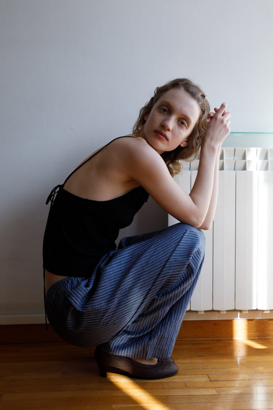 A woman with light skin and wavy hair crouches indoors, calmly gazing at the camera. She wears the Ease Trouser with blue stripes and a black sleeveless top, sunlight streaming across the wooden floor and white wall behind her.