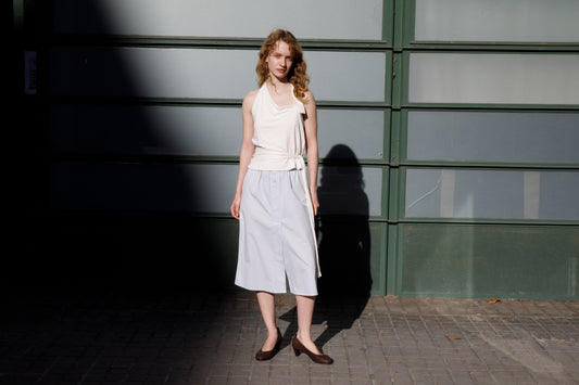 A woman with curly hair stands on a sidewalk by a green and frosted glass wall, wearing a sleeveless white top, the Overlap Skirt - Blue from Deiji Studios, and dark flats as sunlight and shadows fall across the scene.