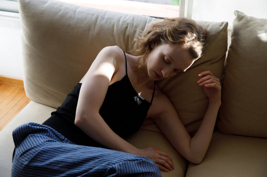 A woman with light brown hair lies sideways on a beige couch, wearing a black tank top and Deiji Studios Linen Ease Trouser - Azure Stripe. Sunlight from a window casts soft shadows across her face and arm.