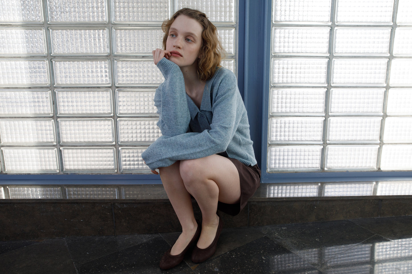 A young woman with light skin and wavy hair sits on a low ledge indoors, looking thoughtful. She wears the Deiji Studios Cross Button Cardigan in Ice Blue, a dark skirt, and brown flats. Glass blocks form the background.