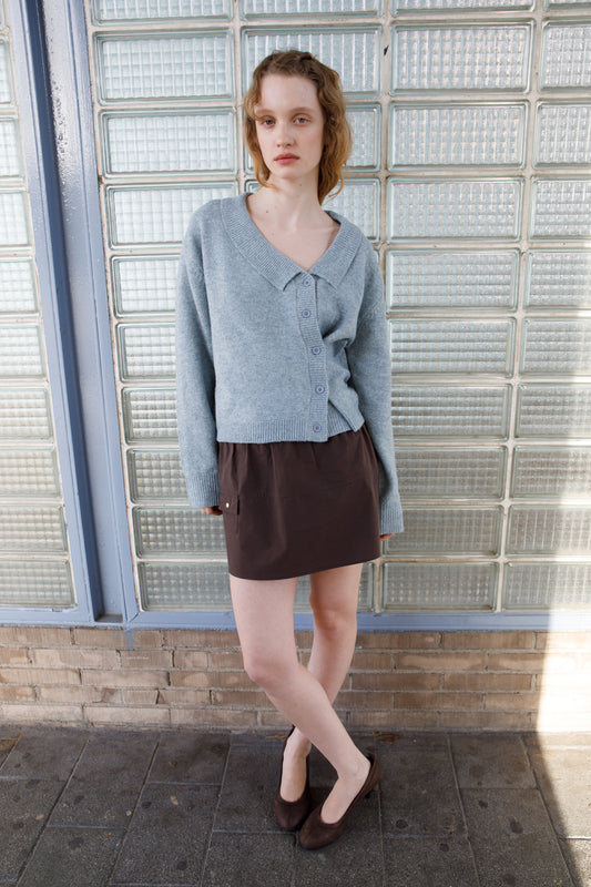 Model wearing the Cross Button Cardigan - Ice Blue, paired with a dark brown mini skirt and brown block-heel mules, standing in front of a glass block wall.