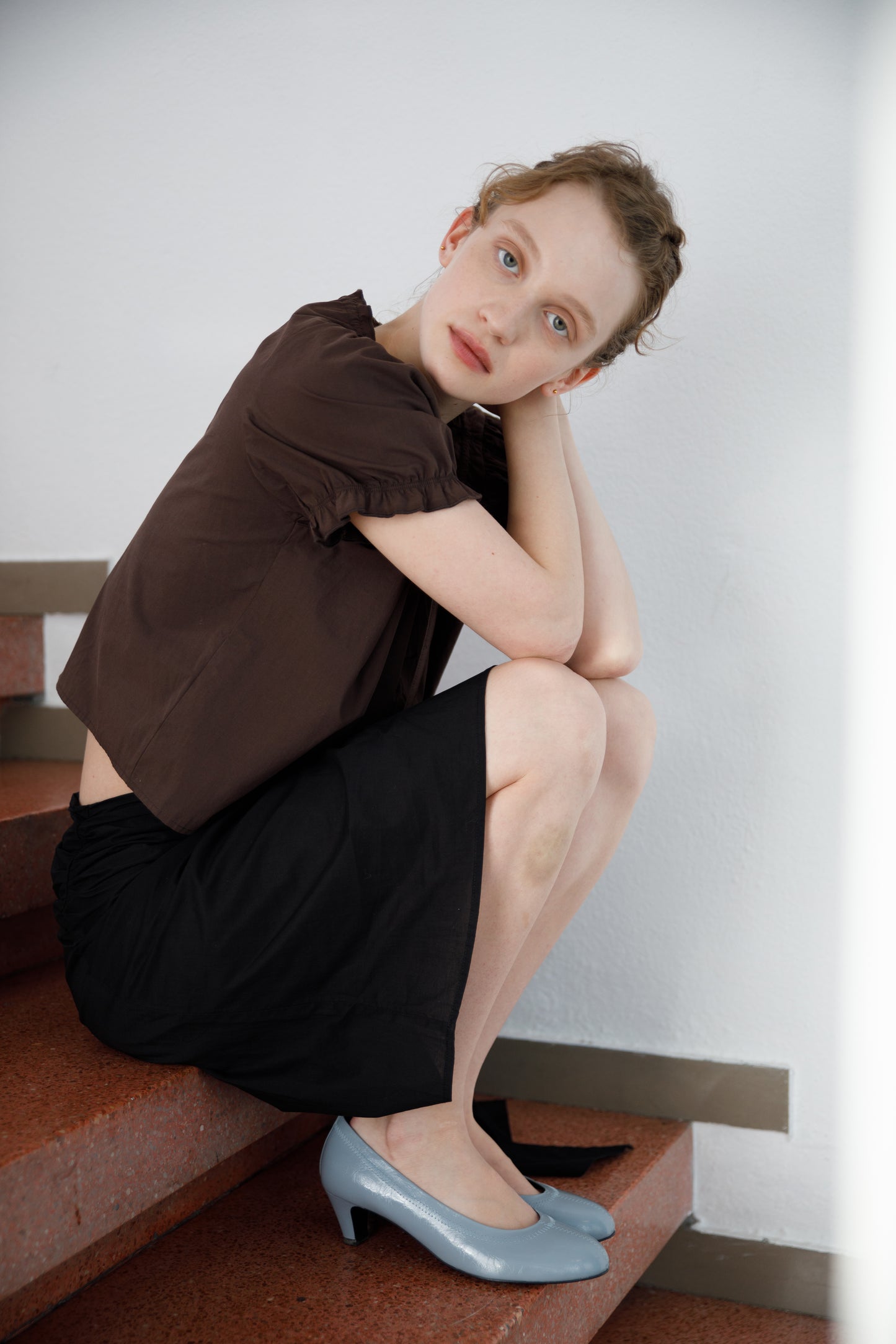 A woman with light skin and curly hair sits on red stairs, resting her arms on her knees. She wears a brown top, the Scarf Midi - Black skirt from Deiji Studios, and light blue heels, looking calmly at the camera.